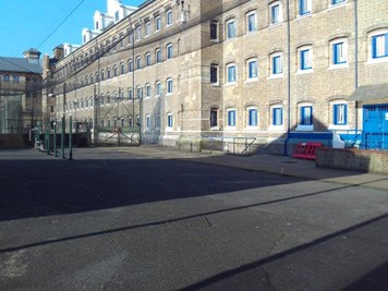 Photograph of the D wing exercise yard, a concreted area with a four-storey brick building to one side. The building has rows of regularly-spaced windows along its length.
