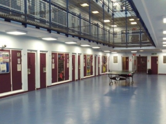 Photograph of the interior of the  ISFL communal area, in which three storeys with balconies on the upper two are visible. The walls on the ground floor are painted white and the cell doors in them are burgundy red. There is a table tennis table in the middle of the space.