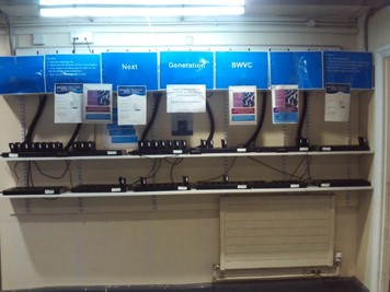 Photograph of a docking station for body-worn cameras, sitting on shelves in an alcove. Instruction signs are fixed above where the cameras are.