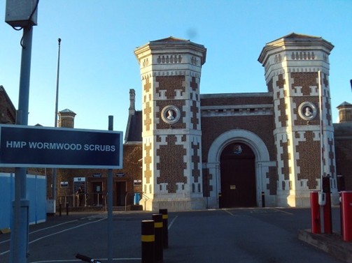 Photograph of the main front gate at Wormwood Scrubs, a large structure with imposing towers on either side. In the foreground there is a sign with the name of the institution on it.