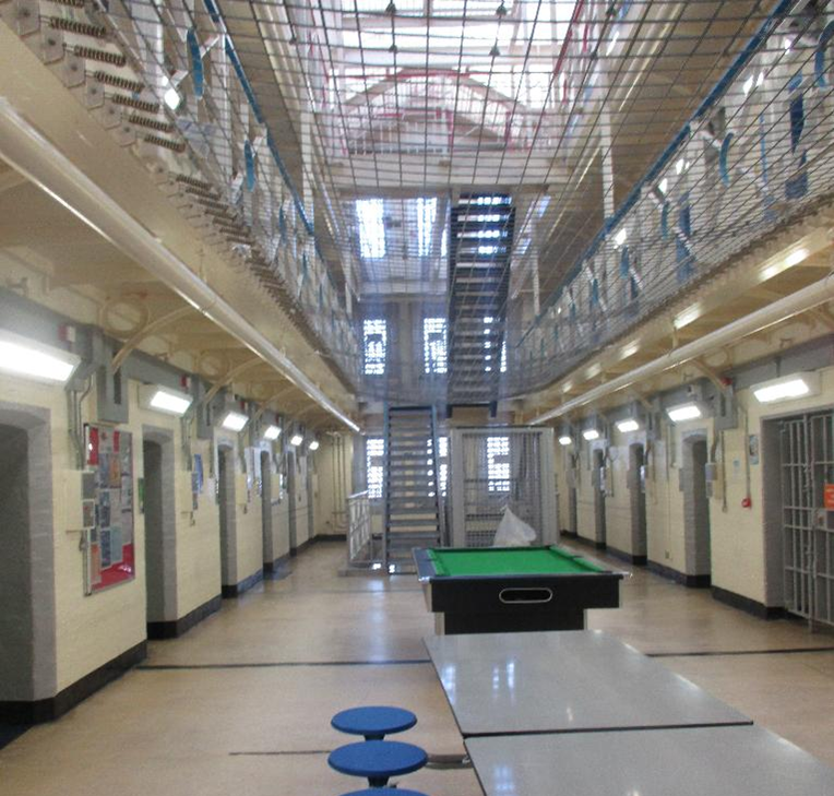 Photograph of an indoor recreational area, showing rows of cells on both sides, a pool table, and a table with attached blue stools in the centre. The space features metal staircases, overhead netting, and barred windows.