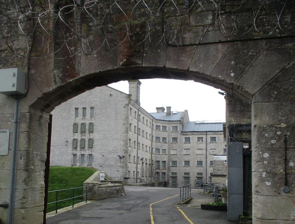 Photograph of a stone archway entrance leading to a tarmacked area surrounded by multi-story, grey stone buildings with barred windows. Yellow lines mark pathways on the ground, with barbed wire visible atop the archway.