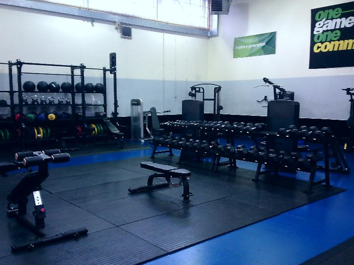 Photograph of an indoor gym area featuring various weightlifting equipment, including benches, dumbbells, and weight plates organized on racks. Walls display motivational banners with phrases "one game one community," and the floor is covered with black rubber mats surrounded by blue flooring.