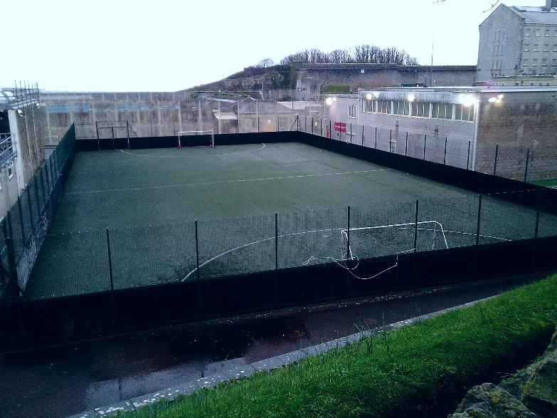 Photograph of an outdoor sports court enclosed by high fences and surrounded by concrete walls and buildings. The court features artificial turf with white boundary lines and a small goalpost.