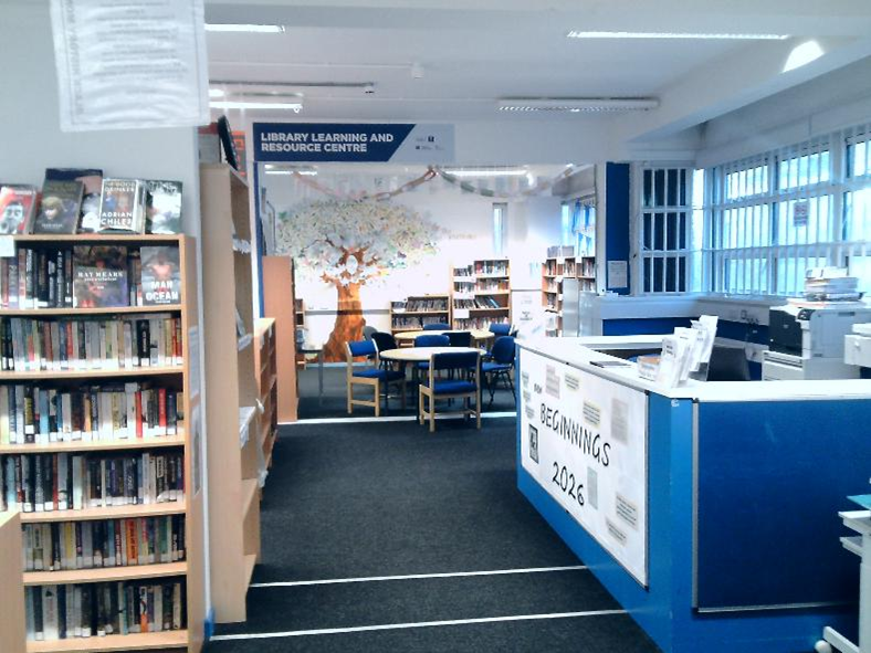 Photograph of a library interior showing bookshelves, a reading area with tables and chairs, and a service desk. The space is well-lit with natural light from large windows, featuring a mural of a tree on the back wall and a sign indicating "Library Learning and Resource Centre."