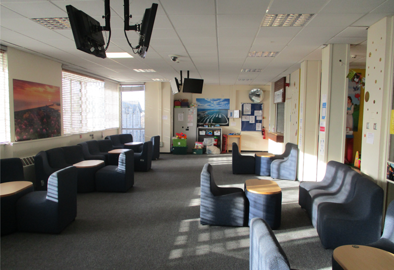 Photograph of a spacious lounge area featuring multiple dark blue cushioned chairs and small round wooden tables arranged for group seating. Natural light enters through large windows on one side, with wall-mounted televisions and colourful artwork enhancing the welcoming atmosphere.