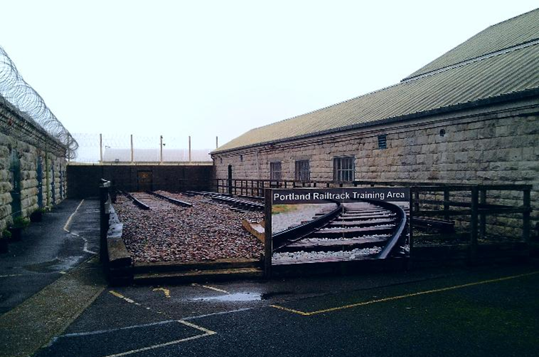 Photograph of an outdoor training area featuring railroad tracks labelled "Portland Railtrack Training Area" on a sign. The scene includes gravel-covered tracks, stone buildings on either side, and a barbed wire fence along the left wall.
