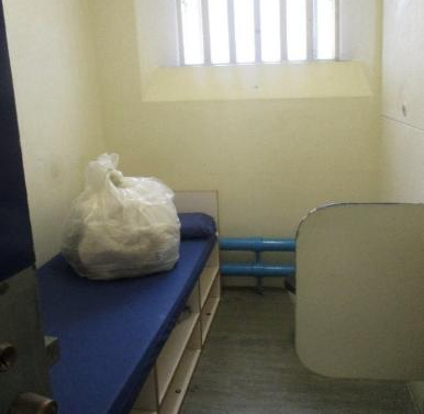 Photograph of a small, sparsely furnished room with a narrow blue mattress on a built-in bed frame and a plastic bag placed on top. The room features a barred window, pale yellow walls, exposed blue pipes along the floor, and a partial white partition on the right side.