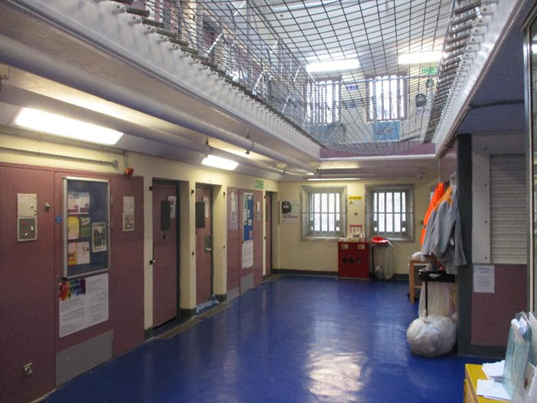 Photograph of an indoor prison corridor showing cell doors along one side and a blue floor. The area includes barred windows, overhead security netting, and various items such as clothing and bags placed near the walls.