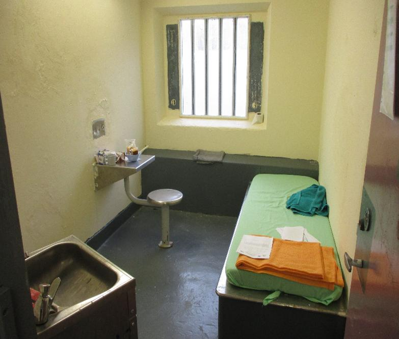 Photograph of a small prison cell featuring a single bed with folded blankets, a metal sink, and a stool attached to a small table. The cell has a barred window, plain walls and minimal furnishings.