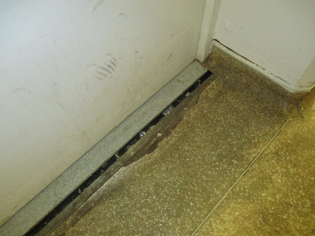 Photograph of damaged flooring on the inpatient unit. The picture focuses on the edge of the room, where the flooring has come away from the wall.