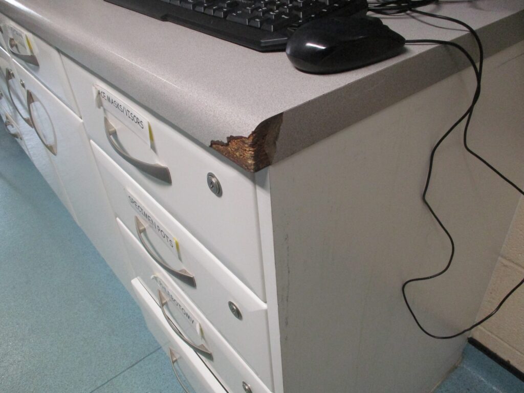 Photograph of a damaged cabinet in the health facility on E wing. The photo focuses on the corner of a unit that apperas to have have been worn or torn away, exposing the plywood underneath.