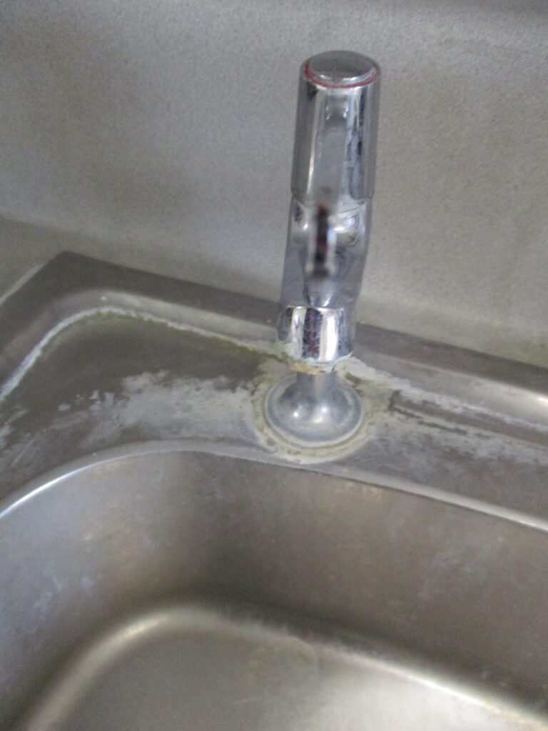 Photograph of a damaged and dirty tap in the health care facility on E wing. The close-up photo shows staining and water damage on and around the vertical part of the tap and the basin around its base.