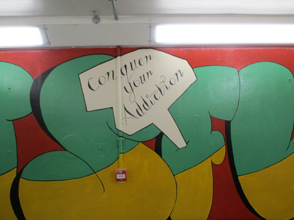 Photograph of a large wall mural on the incentivised substance free living unit. The letters of the ISFL acronym are painted in bright colours as a background, and in a speech bubble in the centre there is the slogan 'conquer your addictions'.