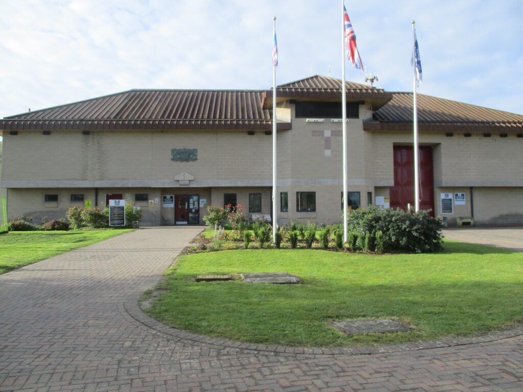 Photograph of the exterior of HMP Bullingdon. There is an area of grass and shrubs, with three flagpoles, in front of a substantial three-storey building. Windows run along the bottom of the building.