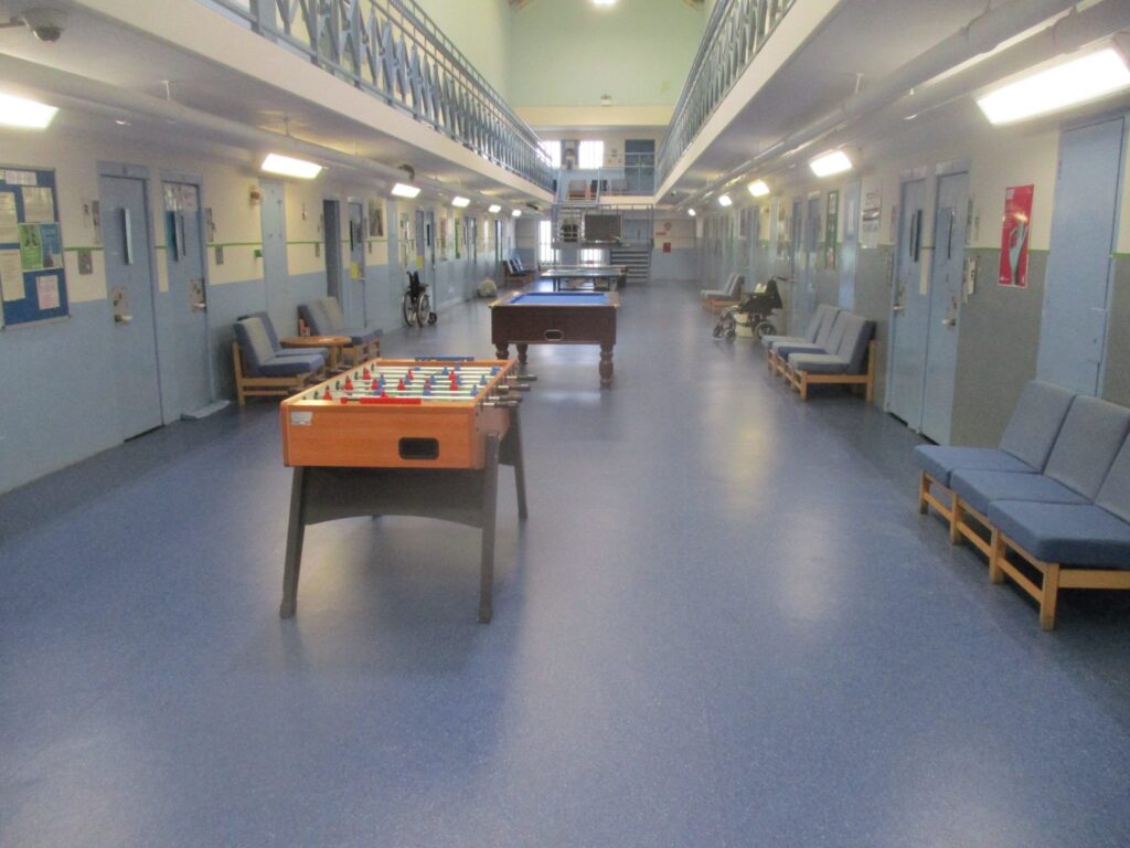 Photograph of a wing communal area. Table football and pool tables are positioned down the middle of the hall and easy chairs are arranged against the walls on either side.