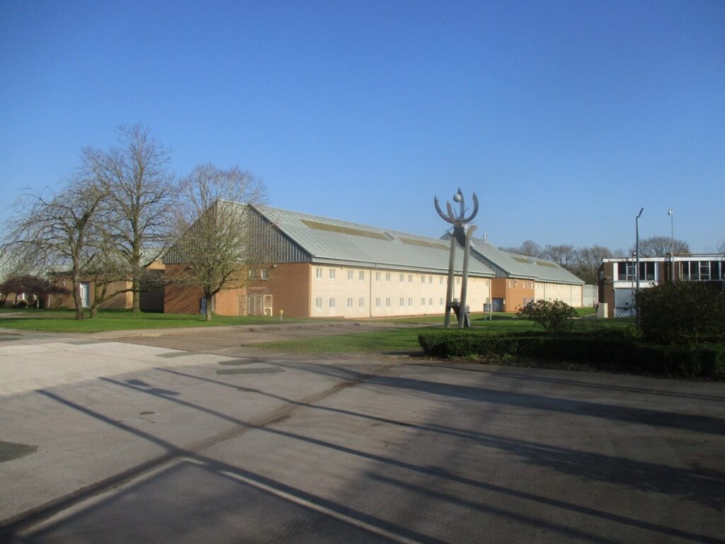 Photograph of the grounds at HMP Stoke Heath, with a mixture of concrete and grass areas at the front of the image. At the back there are a series of substantial two-storey building. There are some trees next to the buildings.