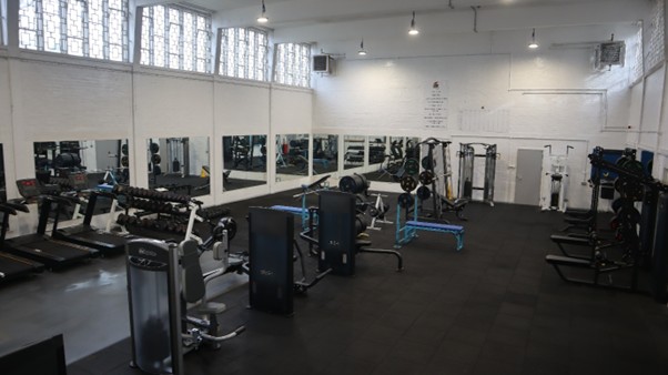 Photograph of the gym after refurbishment The walls are painted white and gym equipment is arranged around the edge of the room and in the centre.