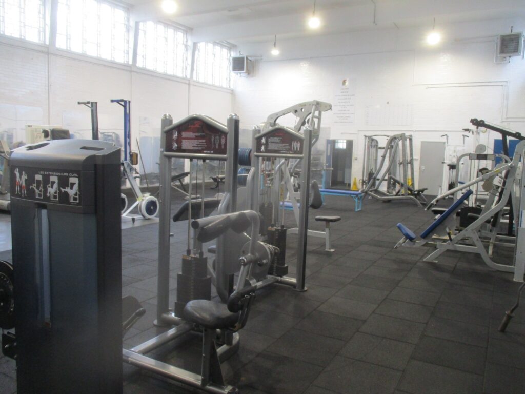 Photograph of the gym before refurbishment. The walls are painted white and the floor is grey. Pieces of exercise equipment are arranged around the edge and in the middle of the room.