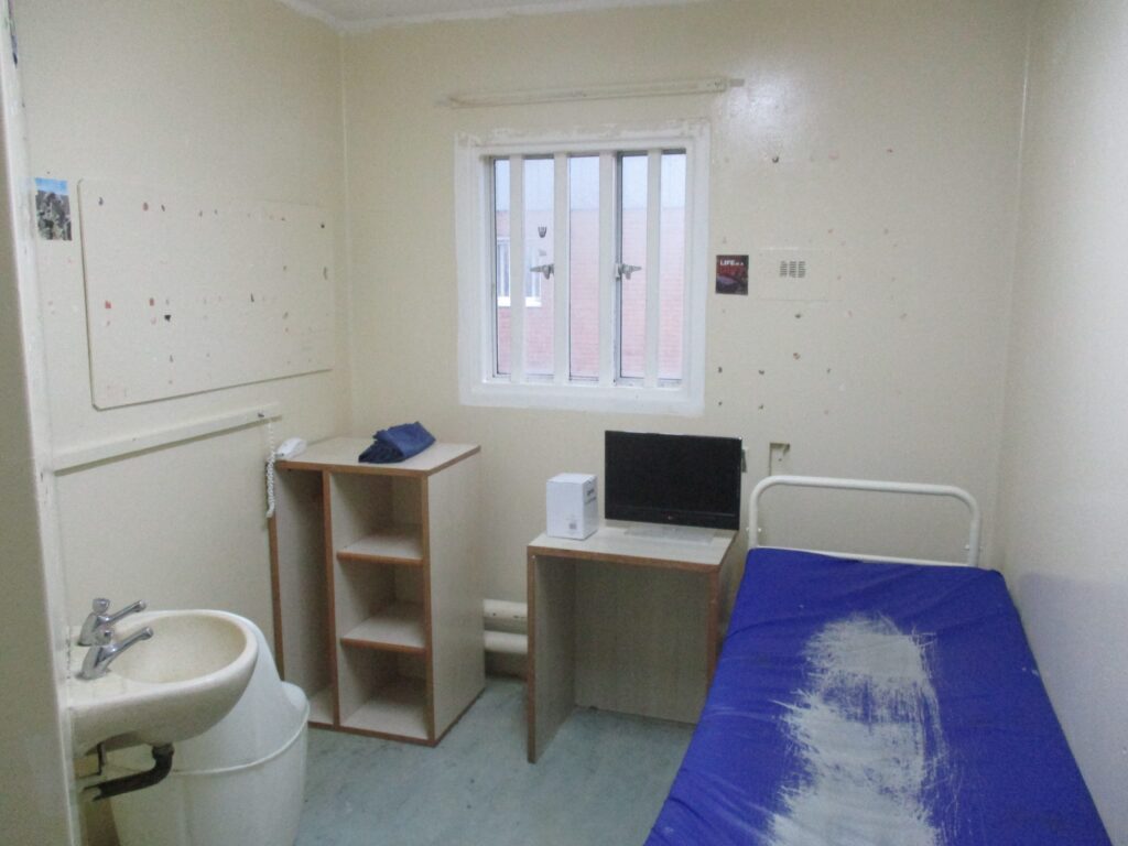 Photograph of an induction unit cell ready for occupation. A bed is positioned to one side of the room and there are two piece of furniture under the window. One of them has a television on top of it. A hand basin can be seen in the nearest corner. The room is painted white.