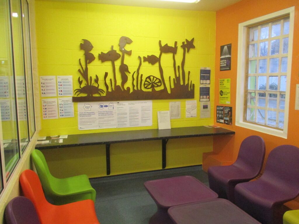 Photograph of the reception holding room, a small space with its walls painted in bright colours and decorated with a mural on the back wall. Chairs are lined up along two sides of the room and there is a coffee table in the middle. A shelf on the back wall has some information leaflets on it and there are further displays of information on the walls.
