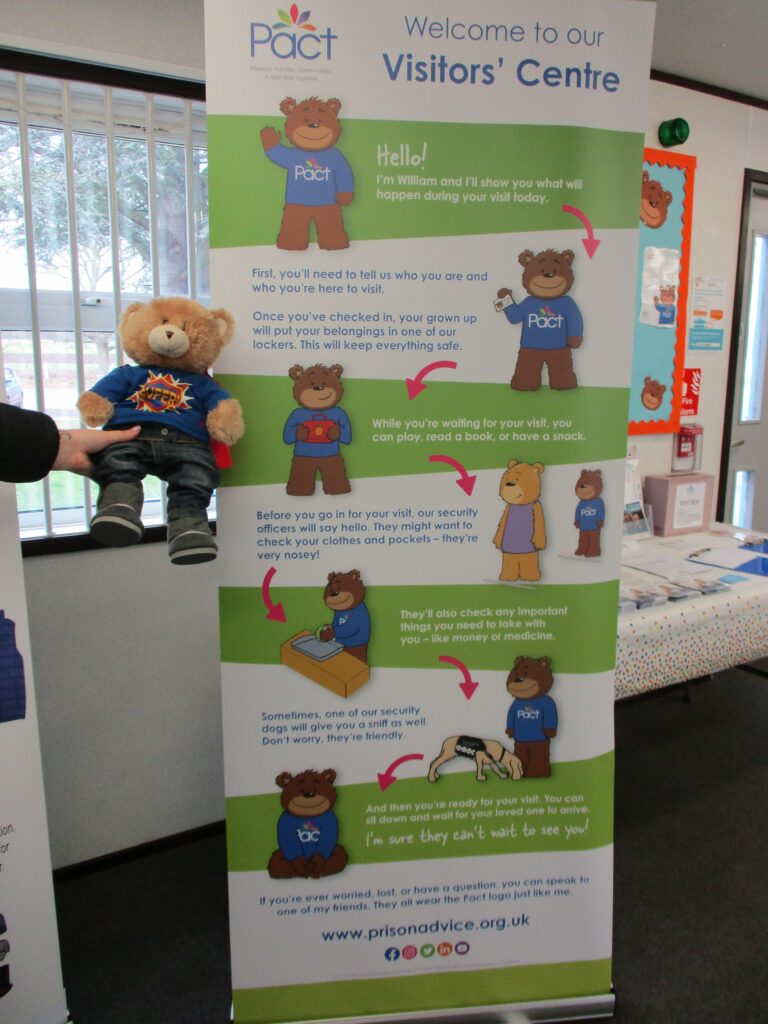 Photograph of the William the Bear display, explaining visits to children. Someone is holding William in one side of the photograph. He is wearing black jeans and boots and a blue jumper.