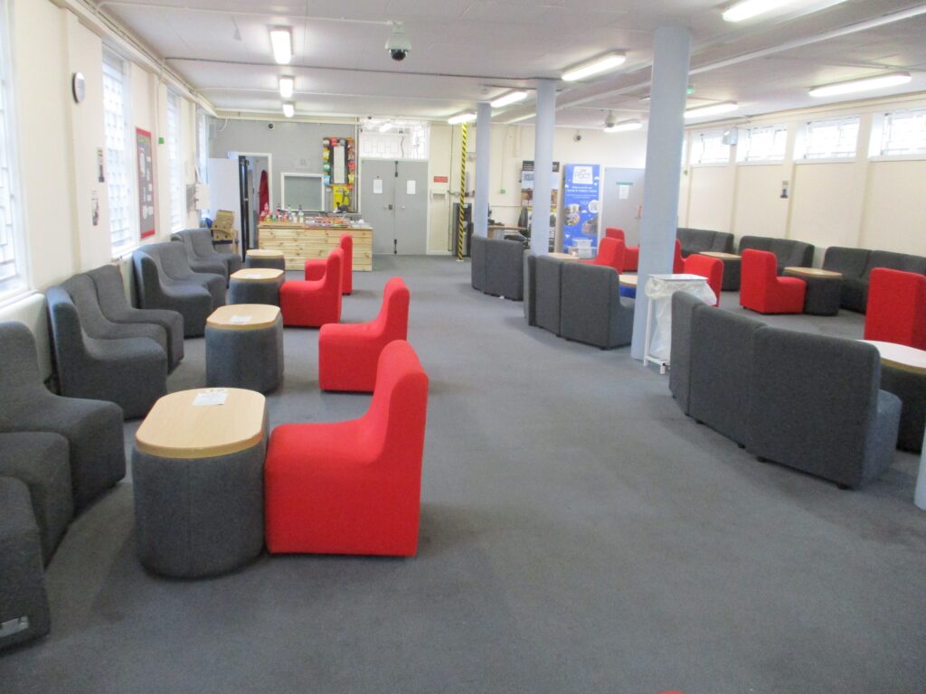 Photo of the visits hall. Seats and tables are arranged down both sides and the centre of the room. The carpet is grey and the seats are upholstered iin red or grey fabric.