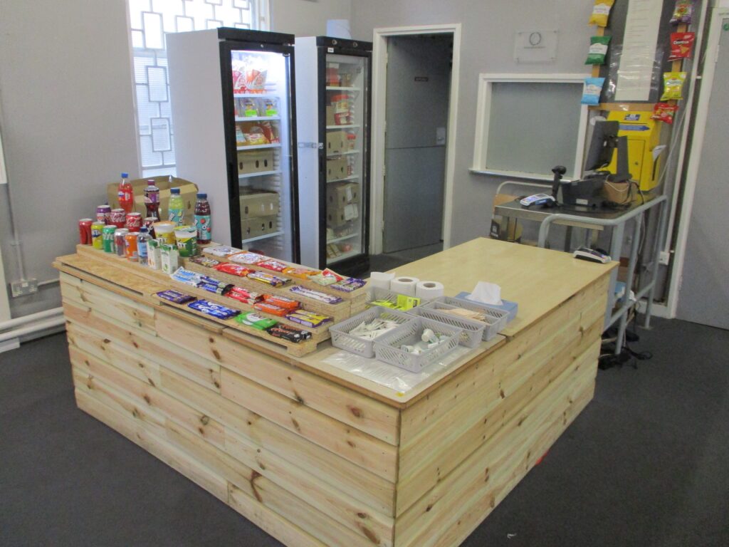 Photograph of the visits hall canteen counter, an L-shaped wooden construction. Behind the counter can be seen two fridges stocked with chilled products.