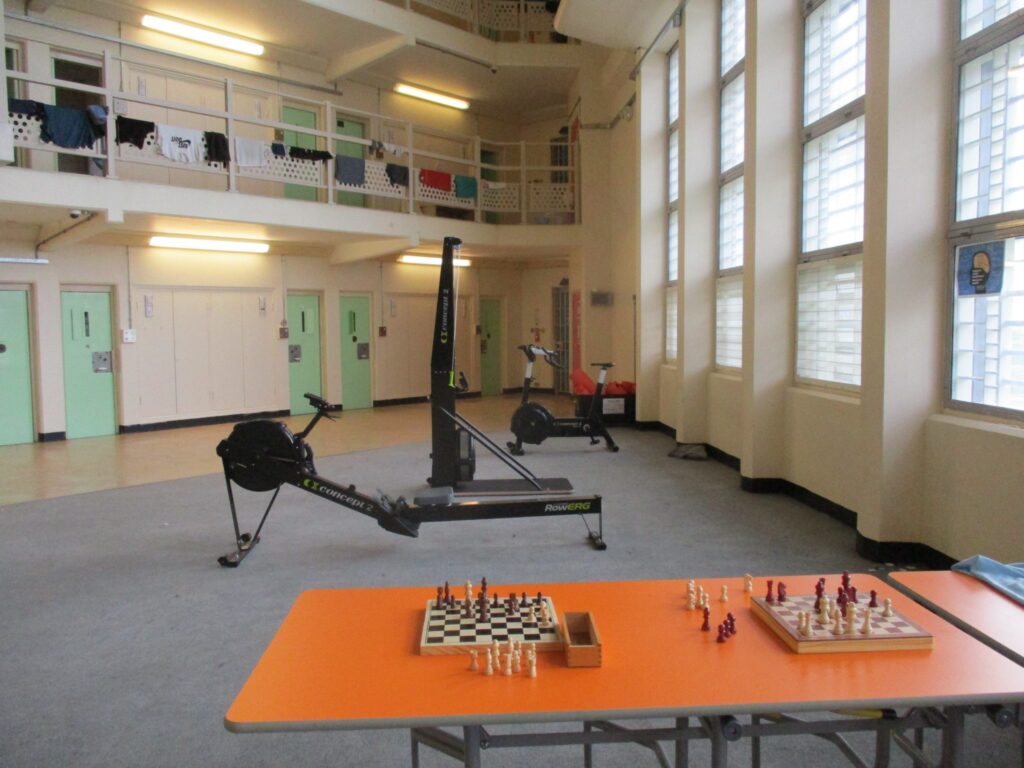 Photograph of the interior of the enhanced wing. It shows a communal. double height space. On the ground level a table with two chess boards on it and some exercise equipment can be seen. Behind that equipment cell doors coloured mint green can be seen in the wall. A balcony can be seen on the first floor at the rear.