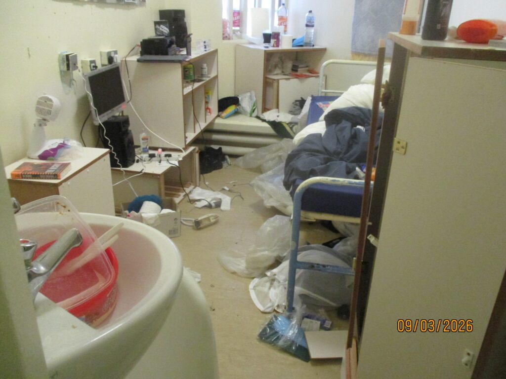 Photograph of a cluttered cell. Personal effects spill off the furniture onto the floor