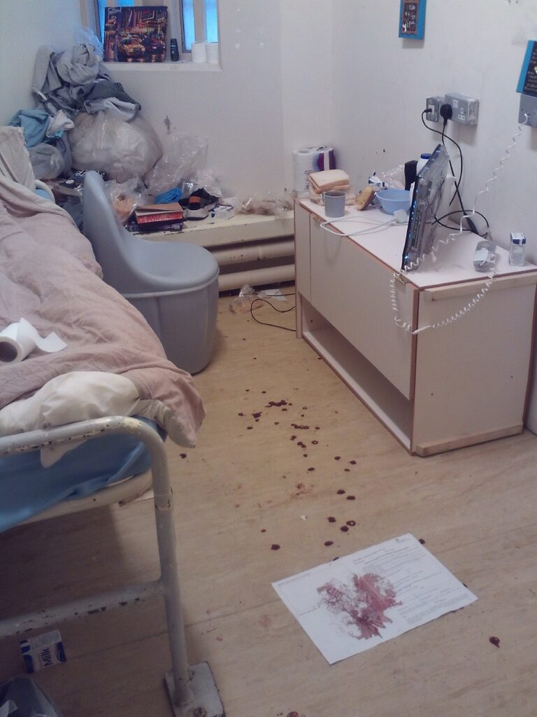 Photograph of a cell that was being used for constant supervision at the time of the inspection. Blood can be seen on the floor and the occupant's content are stowed at the back of the cell.