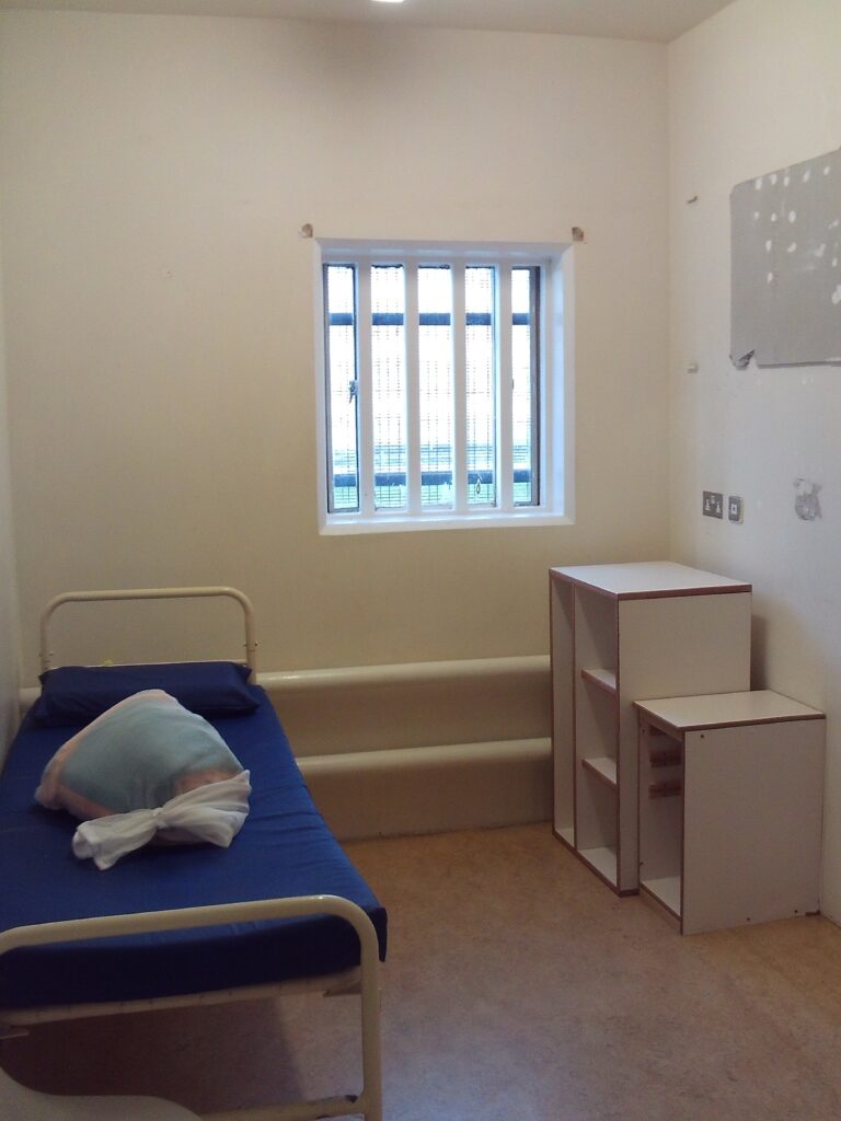 Photograph of a first night cell. The cell has a bed to one side and some furniture to the other. There is a window in the rear wall.
