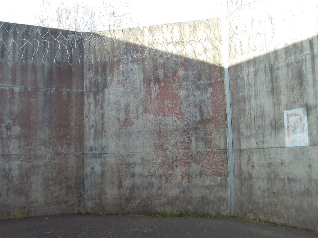 Photograph of the segregation unit exercise yard, a space with concrete walls and ground. There is a basketball hoop on one wall.