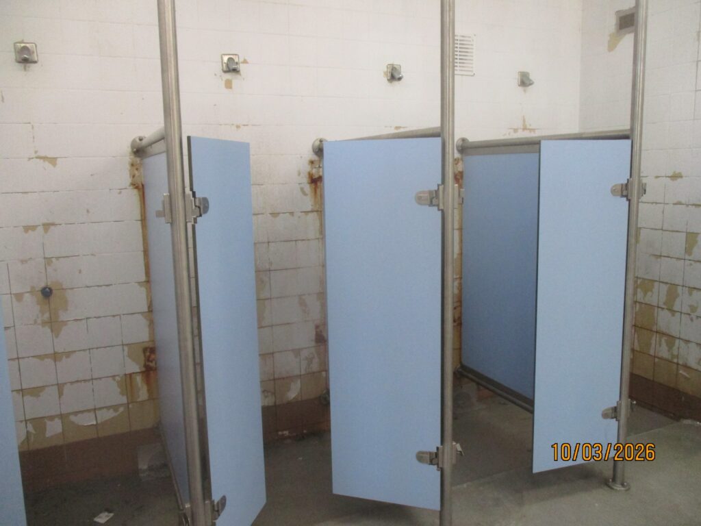 Photograph of a row of shower cubicles. The doors and screens between the showers are light blue in colour and the walls are covered in white tiles.