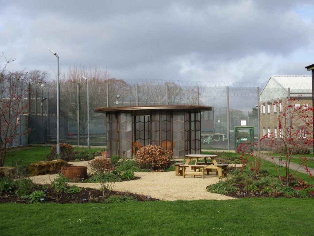 Photograph of an outdoor garden area, featuring a circular glass structure, picnic tables, and landscaped plants. Surrounding elements include tall fences topped with barbed wire, security cameras, and buildings under a cloudy sky.