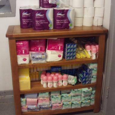 Photograph of a wooden shelf stocked with various hygiene products, including multiple packs of toilet paper, sanitary pads, and deodorants.