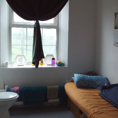 Photograph of a small room featuring a toilet on the left and a single bed with orange bedding and blue pillows on the right. Window with dark curtain partially drawn above radiator with towels, and wall calendar visible on right side.