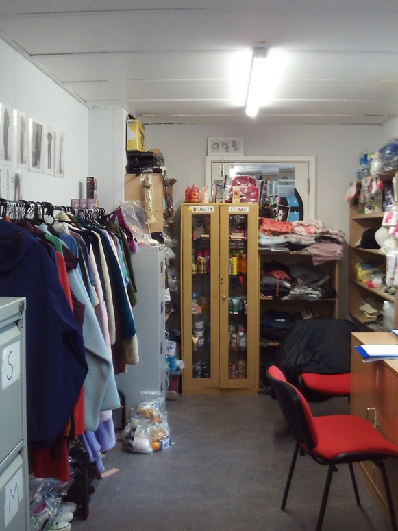 Photograph of a small, cluttered storage room with clothing racks on the left, shelves filled with various items on the right, and a tall cabinet with glass doors labelled 'beauty items'. The room features fluorescent ceiling lighting, a red chair in the foreground, and stacks of folded clothing on shelves and surfaces.