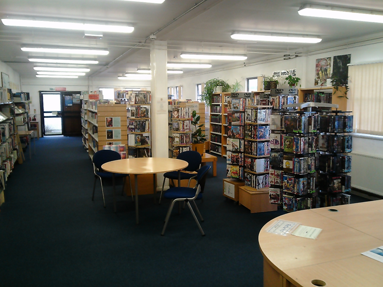 Photograph of a library interior showing rows of bookshelves filled with books and DVDs, along with round tables and chairs for reading or studying. The space is well-lit with overhead fluorescent lights, and plants and posters add decorative elements throughout the room.