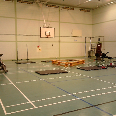 Photograph of an indoor gymnasium setup with exercise equipment and mats arranged on a green court marked with white lines. The scene includes stationary bikes, weights, and wooden platforms under two basketball hoops.