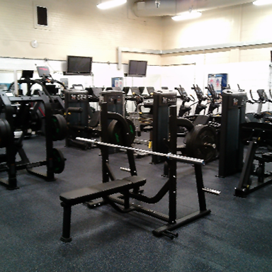 Photograph of an indoor gym featuring various weightlifting and cardio equipment arranged on a dark rubber floor. Key elements include a central bench press with a barbell, multiple weight machines, stationary bikes, and wall-mounted TVs.