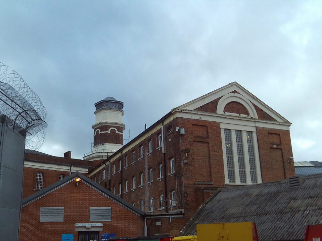Photograph of HMP Winchester, showing a large, old brick building with multiple windows and a tall cylindrical tower. Barbed wire lines the top of a nearby wall. The sky is overcast.
