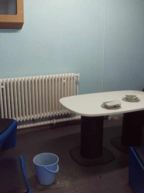 Photograph of a bucket catching a leak in a reception holding room. The room has light blue walls, a white table surrounded by blue and black chairs and a white radiator is mounted on the wall behind the table.