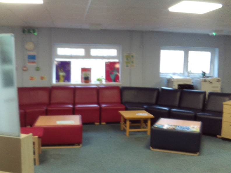 Photograph of an induction room, with two sofas, one red and one black, arranged around three wooden tables. The room has light blue carpet and large windows, some windows have posters on.