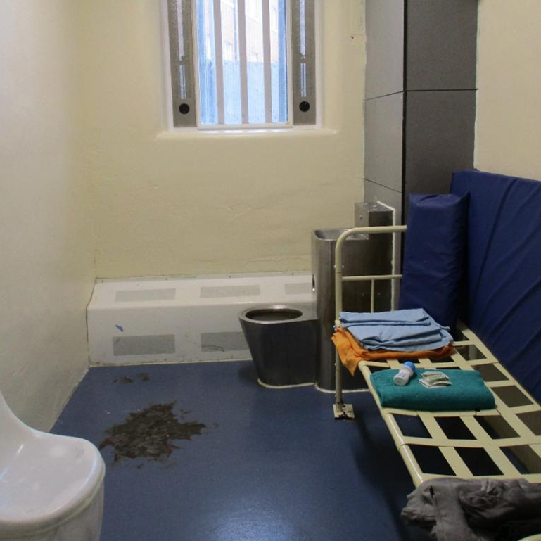 Photograph of a segregation cell, with a barred window, metal bed frame with folded items on top, and a blue mattress and cushion leaning against the wall behind the bed frame. There is a metal toilet and sink on the window wall. The cell has plain cream walls, a blue floor with a dark stain near the toilet, and a white plastic chair.