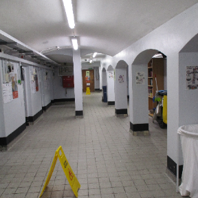 Photograph of the segregation unit, with a long, tiled walkway with white walls and arched doorways, showing fluorescent ceiling lights and safety signs on the floor. The hallway contains various posters on walls, a trash bin on the right, and cleaning equipment visible through one archway.