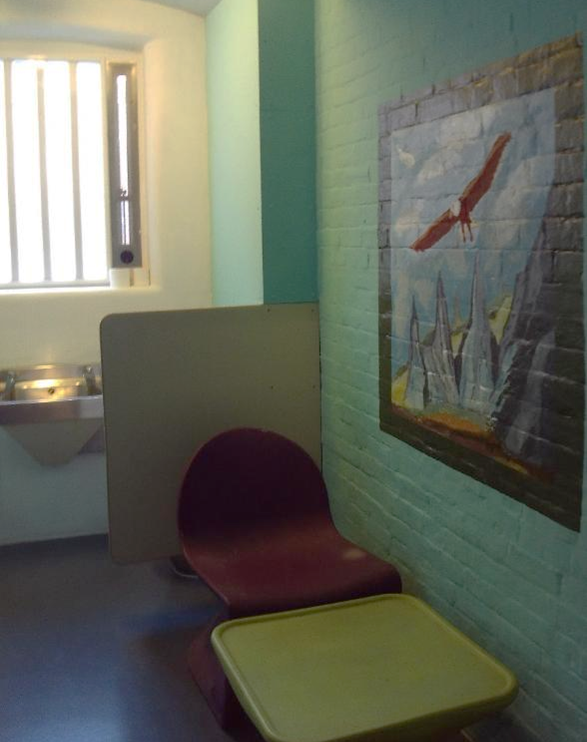 Photograph of the Listener suite, showing a small room with a barred window, a purple chair, and a green table. A colourful mural painted on a light blue brick wall shows a bird flying over rocks.