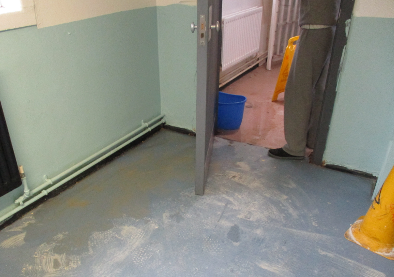Photograph of a leak on Westhill, showing a light blue floor covered in dust and water and an open door leading to another area which is also covered with water and has a blue bucket and a yellow caution sign. Walls are painted light green.