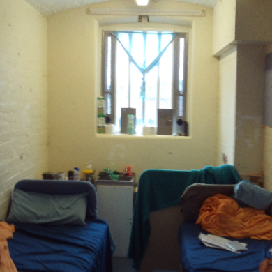 Photograph of a crowded double cell, showing two single beds on opposite sides, each covered with blankets and pillows. A window with bars and a curtain is centred on the back wall above a small sink and unit holding various personal items.