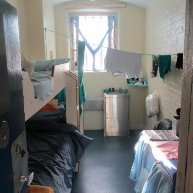 Photograph of a crowded double cell, with bunk beds on the left hand side, a metal sink, and clothes hanging on a line near a barred window. The cell has pale walls, a dark floor, and minimal personal items.